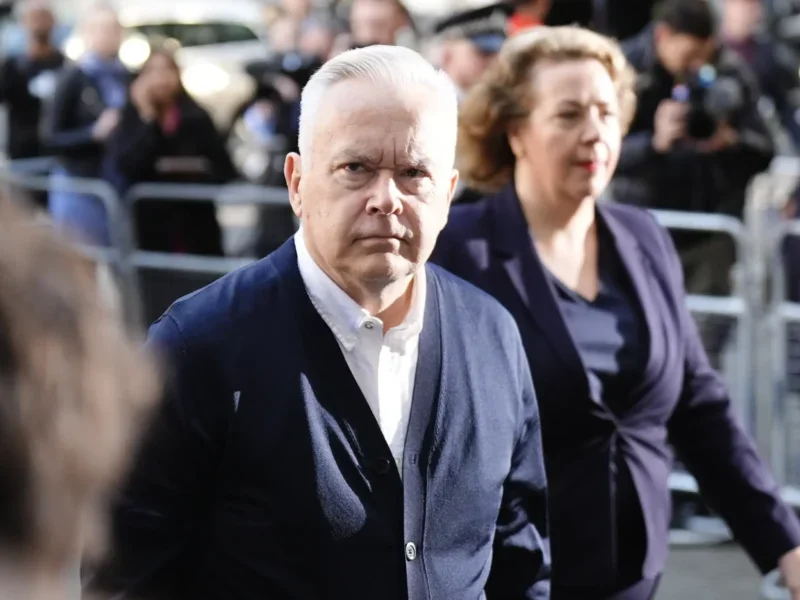 Huw Edwards walking, looking to his right where the camera is and frowning. He's wearing a cardigan and white shirt. Behind him there is a metal fence containing crowds of people.