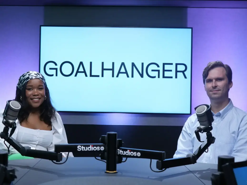 Nicole Logan and Andy Neilson of Goalhanger Podcasts sit in a podcast studio with microphones in front of them and a big screen saying Goalhanger behind them